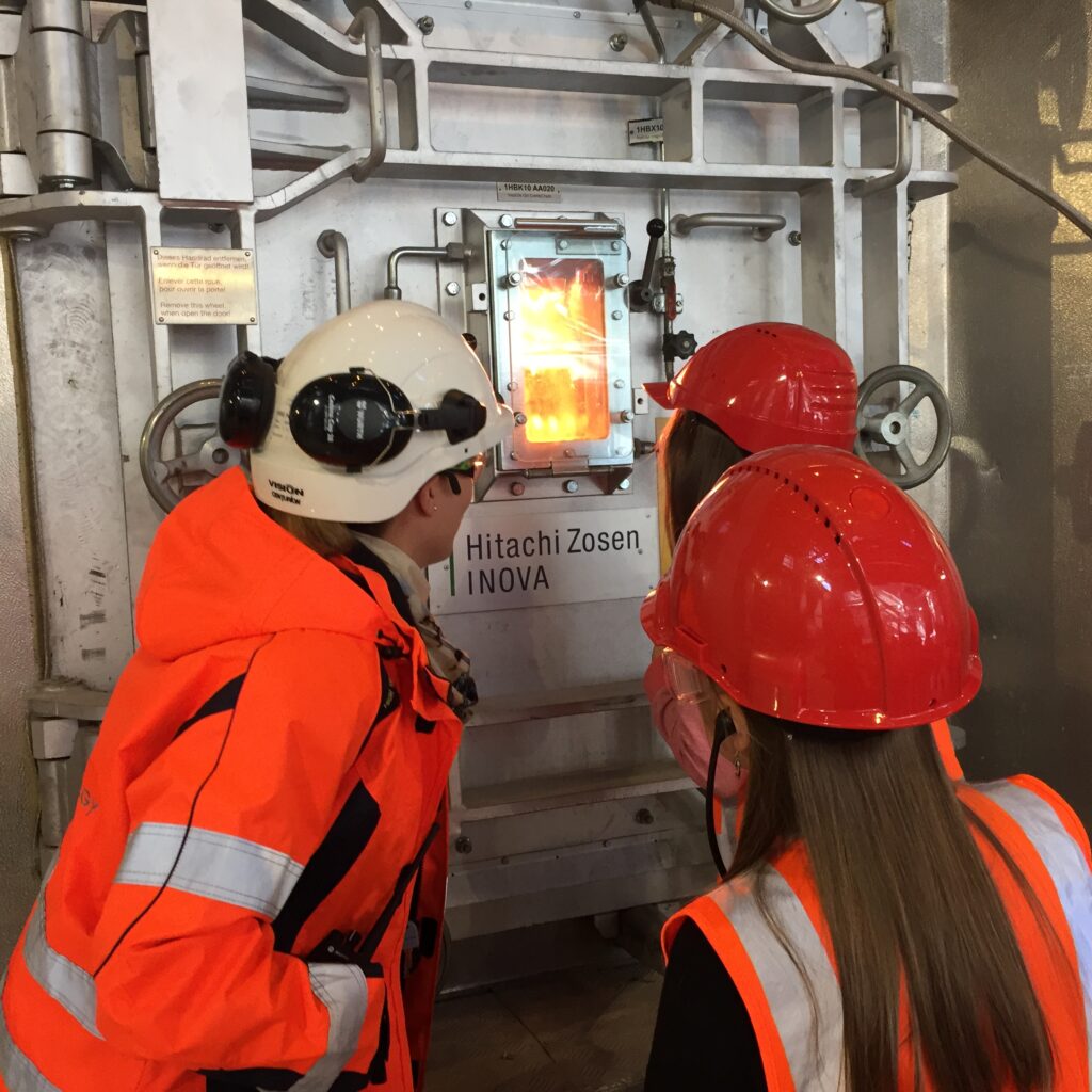 Kolme ihmistä katsoo arinan luukusta. Luukusta näkyy lieskat.